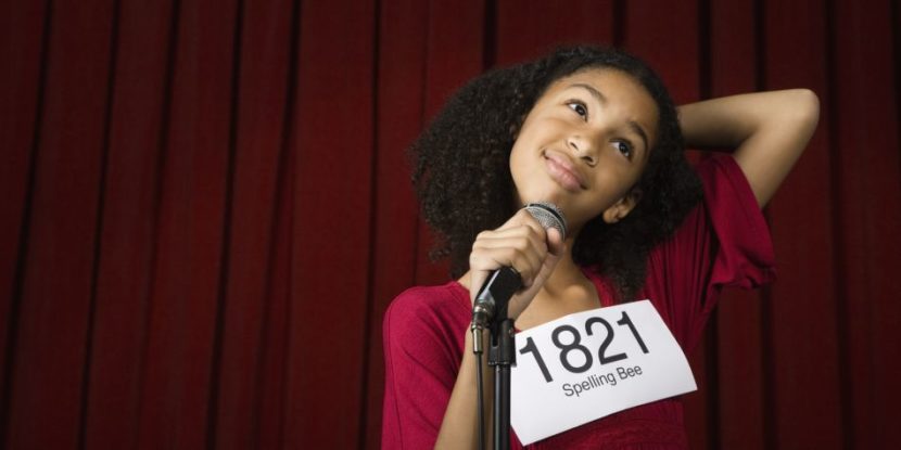A spelling bee contestant (with light-medium skin and coily hair) wears a contestant number and scratches head while standing behind the microphone.