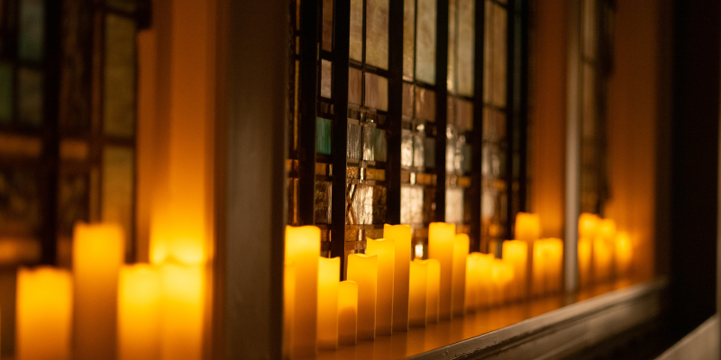 Several lit candles line up against the stained glass window of a dark room
