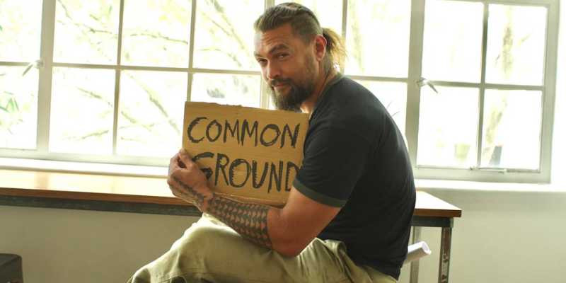 Jason Momoa holding a cardboard sign that says "Common Ground" written with bold black text.