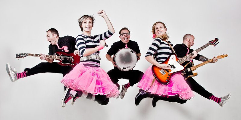The Not-Its! band jump mid-air in front of a white background. Each member is holding a different instrument and dressed in black and pink clothes.