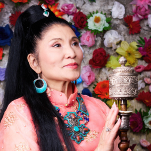 Yungchen Lhamo stands in front of a wall of colorful flowers wearing traditional Tibetian attire and holding a wooden instrument.