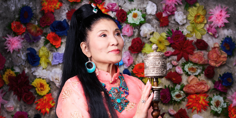 Yungchen Lhamo stands in front of a wall of colorful flowers wearing traditional Tibetian attire and holding a wooden instrument.