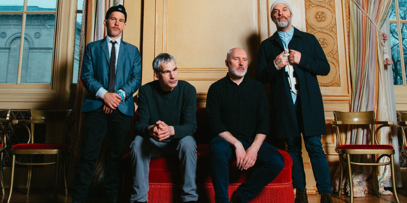 All 4 members of The Bad Plus wearing dark clothing and posing on a red sofa