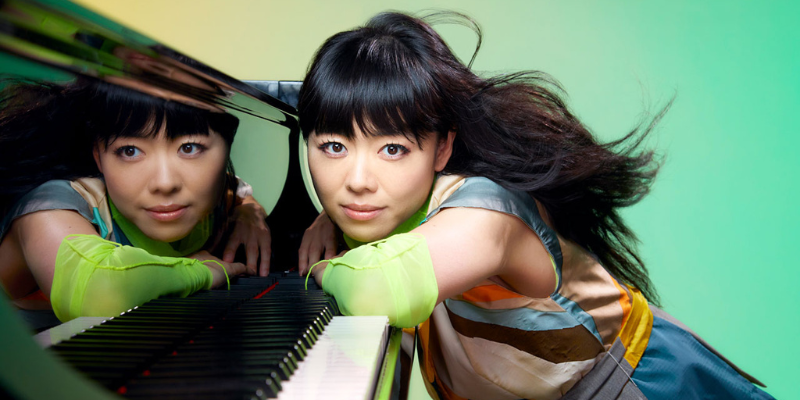 Hiromi (with long black hair and fair skin) smiling and leaning against a grand piano