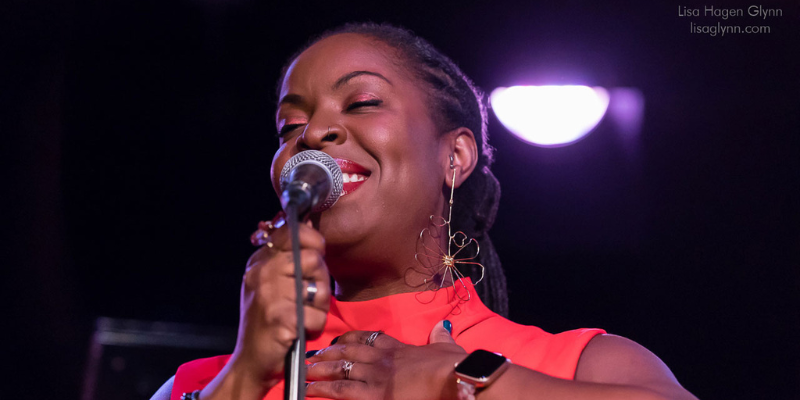 Close-up shot of Johnaye Kendrick (with dark skin, red lipstick, and black braids in a ponytail) singing into the microphone