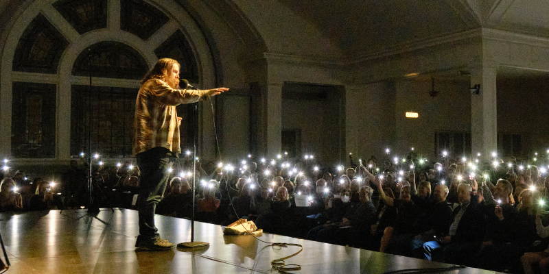 A GrandSLAM presenter speaking onstage to a full crowd of people in The Great Hall holding their phones up as flashlights.