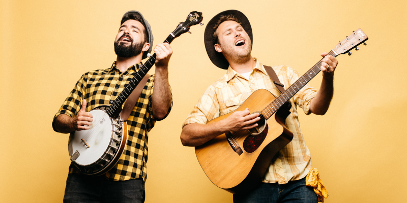 The Okee Dokee brothers playing the banjo and acoustic guitar in front of a yellow background.
