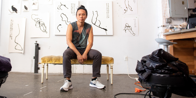 Paul Chan (with black hair in a bun) sitting on a bench and posing in front of a wall of art