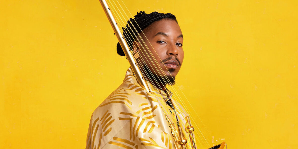 Chief Adujah holding a gold stringed instrument and standing in front of a yellow background.