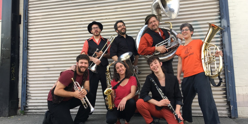 All 7 members of Shpilkis posing dressed in black, orange, and red. Each member is holding a different brass/woodwind instrument.