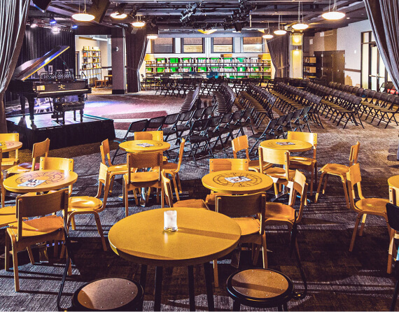 Photo of a mid-sized venue with a stage and carpeted seating area with row seating plus round tables and chairs