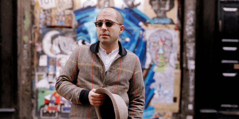 Mahan Esfahani (with light skin, buzzcut, and sunglasses) stands in front of a wall covered with graffiti, holding a fedora.