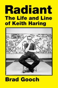 Book cover: A yellow background with an image of Keith Haring sitting in front of his artwork. The image has a thin black outline. Big black text above the picture of Keith says “Radiant, The Life and Line of Keith Haring.”
