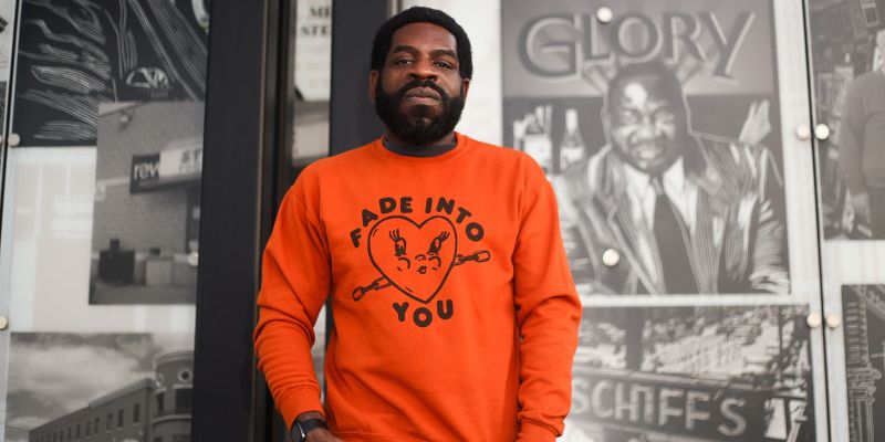 Hanif Abdurraqib (with dark skin, short coily hair, beard, and orange sweatshirt) leans against a wall of old posters.