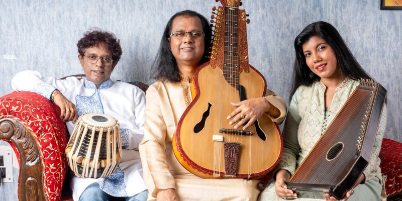 Debashish Bhattacharya poses with a Hindustani slide guitar on a red chaise lounge with two other people, holding different instruments.