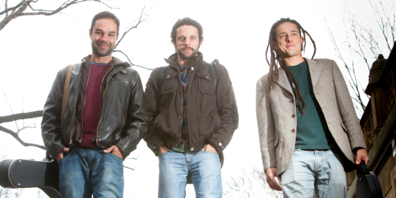 Outdoor group picture of Trio Brasileiro, with the members on the left/right carrying guitar cases