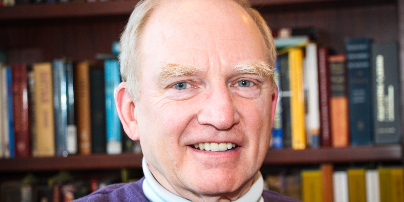 Headshot of Roberyt W. Merry (with fair skin & hair) in front of a background of full bookshelves