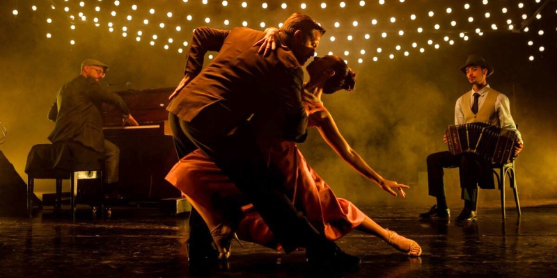 Two dancers perform a dramatic tango move on a stage, spotlighted under a canopy of glowing string lights. A pianist and a bandoneon player accompany them.