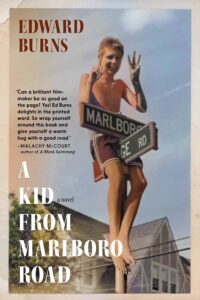 Shirtless young boy sitting atop a street sign labeled "Marlboro Rd" on the cover of the book "A Kid From Marlboro Road" by Edward Burns. The boy smiles and holds three fingers up with one hand and one finger with the other. Critical acclaims from Malachy McCourt is on the left side.