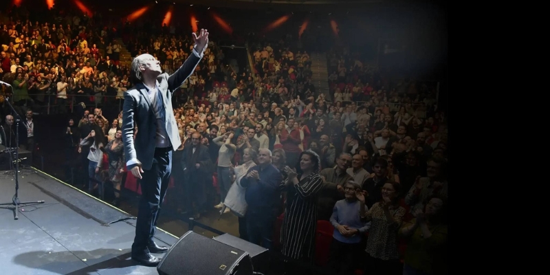 Carlos Nunez stands in the spotlight on stage, gesturing with one hand upwards to a full audience of people.