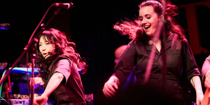 Two people with long hair dance on a stage lit pink, wearing matching black utility jumpsuits. Each person dances behind a microphone.