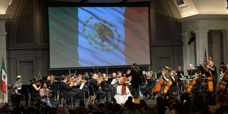 Orquesta NW sits with their instruments on stage as a singer in a white dress performs. The Mexican flag is on a large projector in the background.