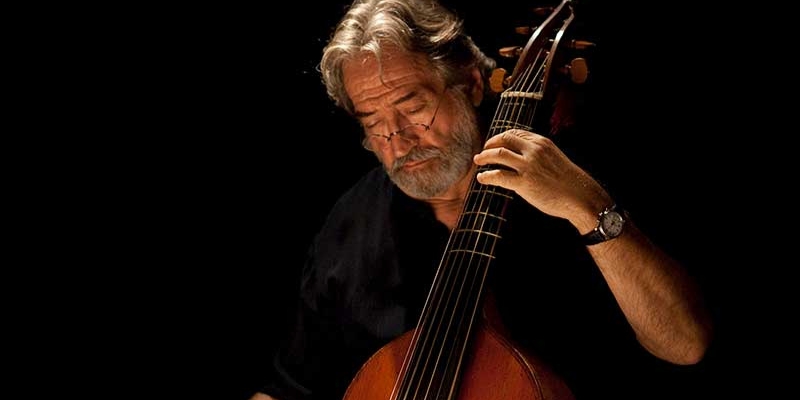 Jordi Savall plays the viola da gamba in front of a black background, wearing black clothing.