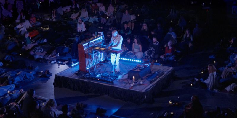 Musician playing synthesizers on a dimly lit stage surrounded by a seated audience in a dark, ambient setting.