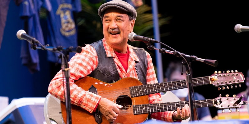 A person wearing a red gingham button-down and newsboy cap smiles and plays a double neck acoustic guitar in front of a microphone.