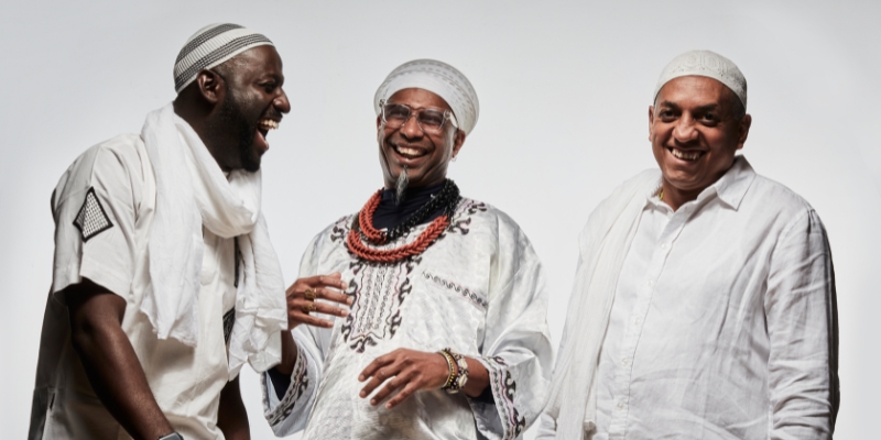 Group photo of Omar Sosa, Seckou Keita, and Gustavo Ovalles. The trio poses in front of a white background, dressed in primarily white cultural attire.