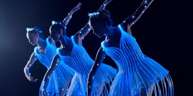 Three ballet dancers in a row pose in a dark room. They wear tutu dresses and tiaras that are lit up by blue lights.