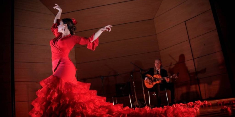 A flamenco dancer wearing a ruffled red dress performs on stage in front of a Spanish guitar player.