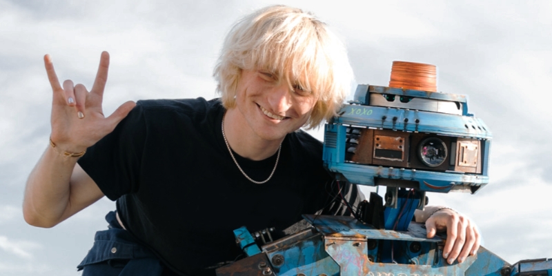 Bailey Ambrose Heller (with fair skin/short blonde hair) poses outdoors and smiles with his arm around a blue robot.