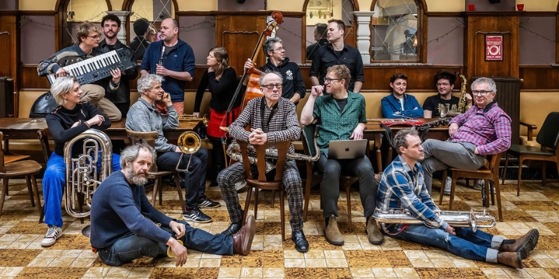 Candid group photo of the Flat Earth Society Orchestra posing at restaurant tables with members holding various instruments. There are 15 members in the photo.