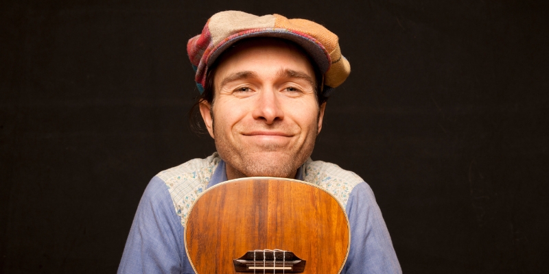 Keeth Apgar from The Harmonica Pocket smiles in front of a black background, holding a ukulele bottom-side up and resting his chin on it.