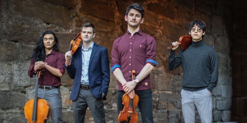 All 4 members of the Kodak Quartet pose in front a stone wall, posing with their stringed instruments.