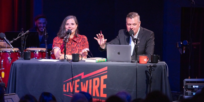 Two people sit at a table with the Live Wire Radio logo and speak into microphones.