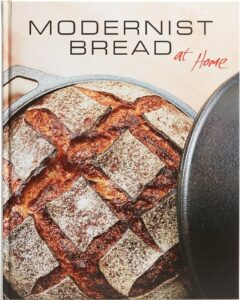 Cover of the book 'Modernist Bread at Home' showing a round loaf of bread with a crosshatch pattern on the crust, partially covered by a cast iron pot lid.