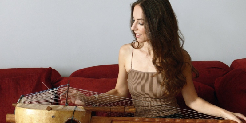 Sophie Lukacs (with fair skin and long brown hair) sits on a red couch and looks down on the kora instrument in her lap, smiling.