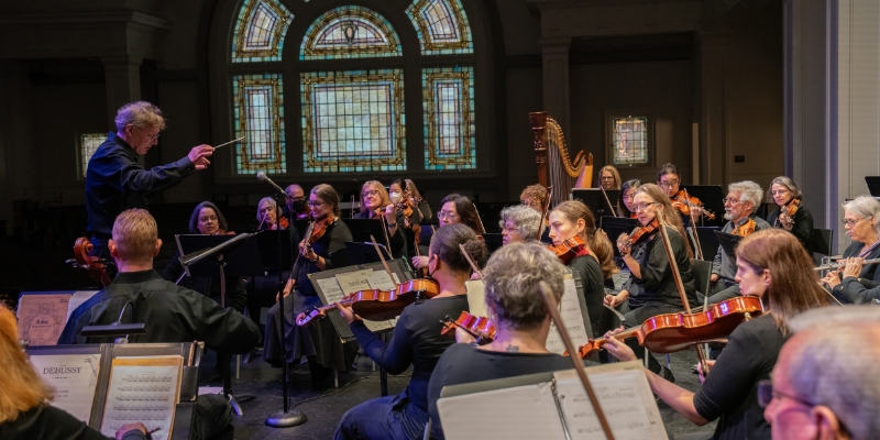 Side shot of Thalia Symphony Orchestra performing on The Great Hall stage, with a stained glass window in the background.