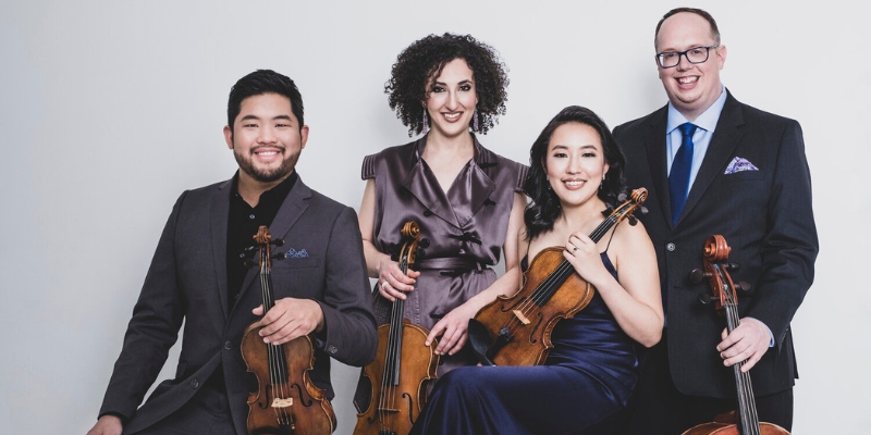 Group photo of the Verona Quartet dressed in formal attire, holding their stringed instruments and posing in front of a white background.