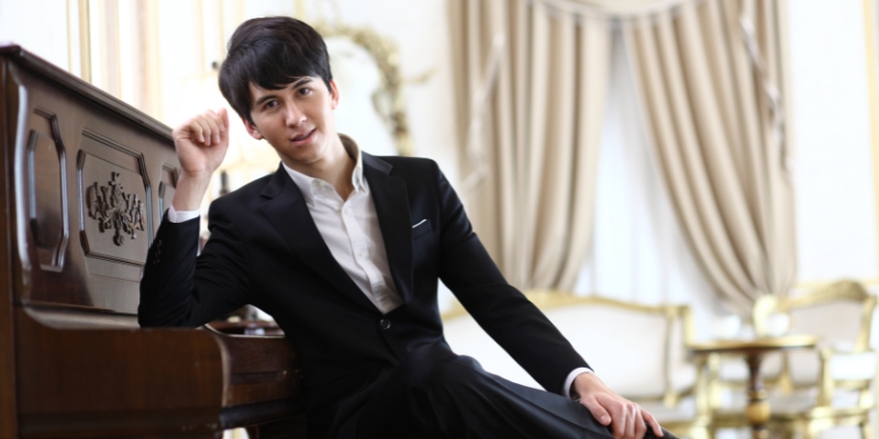 Charlie Albright, wearing a suit, poses on a bench and leans on a baby grand piano with one elbow.