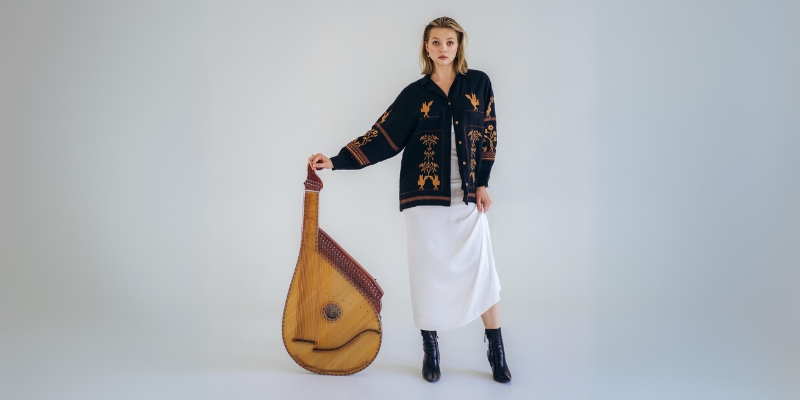 Maryna Krut (wearing a dark patterned cardigan and white skirt) poses and leans with one hand on a bandura instrument.