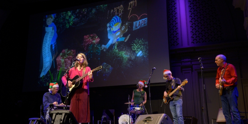 Band performing on the Great Hall stage with Christmas hats, featuring a singer with an acoustic guitar, drummer, keyboardist, bassist, and guitarist, with holiday-themed visuals projected in the background.