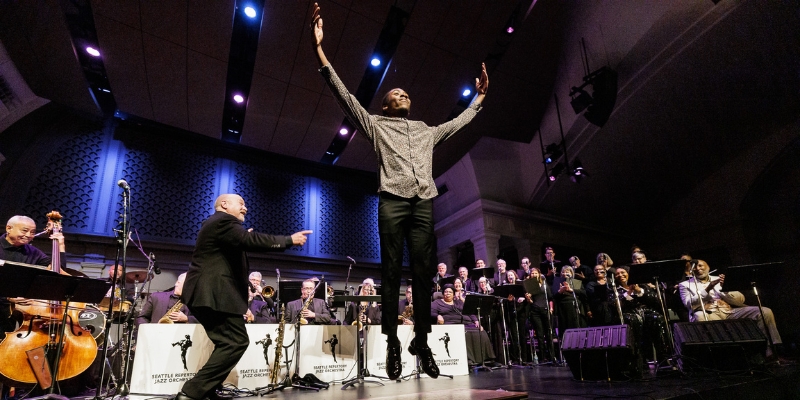 A jazz performance on the Great Hall stage with a tap dancing soloist leaping in mid-air, arms raised, as the conductor gestures toward the dancer. The Seattle Repertory Jazz Orchestra plays behind them with blue stage lighting.