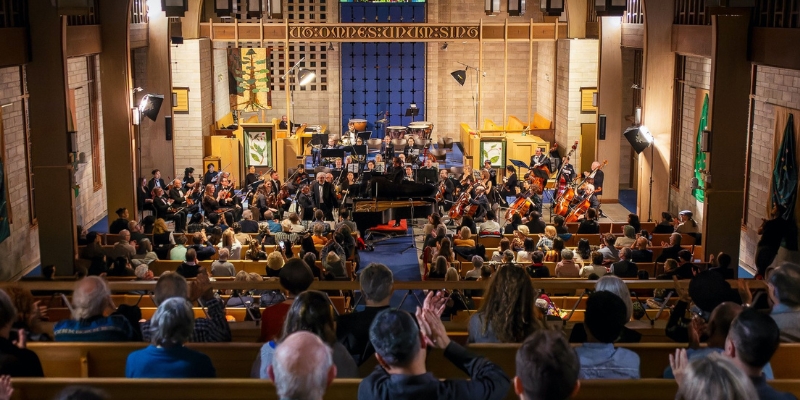 Orchestra performing for a seated audience in a warmly lit church, with musicians playing string and brass instruments on stage under a decorative banner.