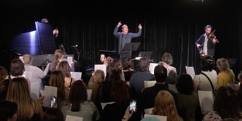 A person conducts on a stage, with a violin player and a filmer using a smartphone. The audience is holding sheet music and singing.