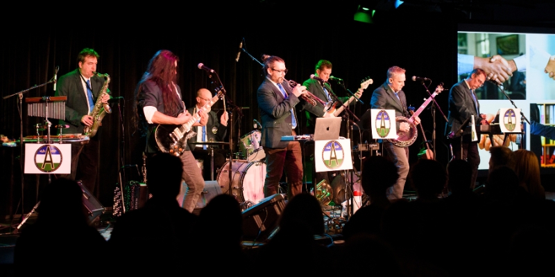 "Awesome" performs on a stage with various instruments, including a saxophone, guitar, trumpet, windchimes, and banjo in front of an audience. The "Awesome" band logo is posted on various instrument stands. All 7 members are wearing dark suits with blue ties.