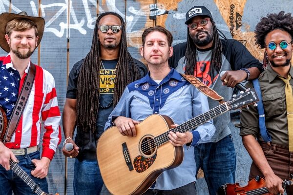 All 5 members of Gangstagrass posing together with instruments, including a banjo, guitar, and fiddle, against a graffiti-covered wooden backdrop.