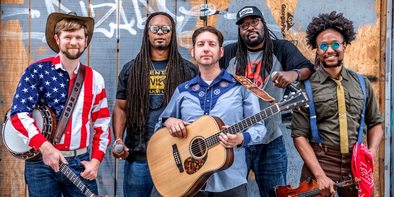 All 5 members of Gangstagrass posing together with instruments, including a banjo, guitar, and fiddle, against a graffiti-covered wooden backdrop.